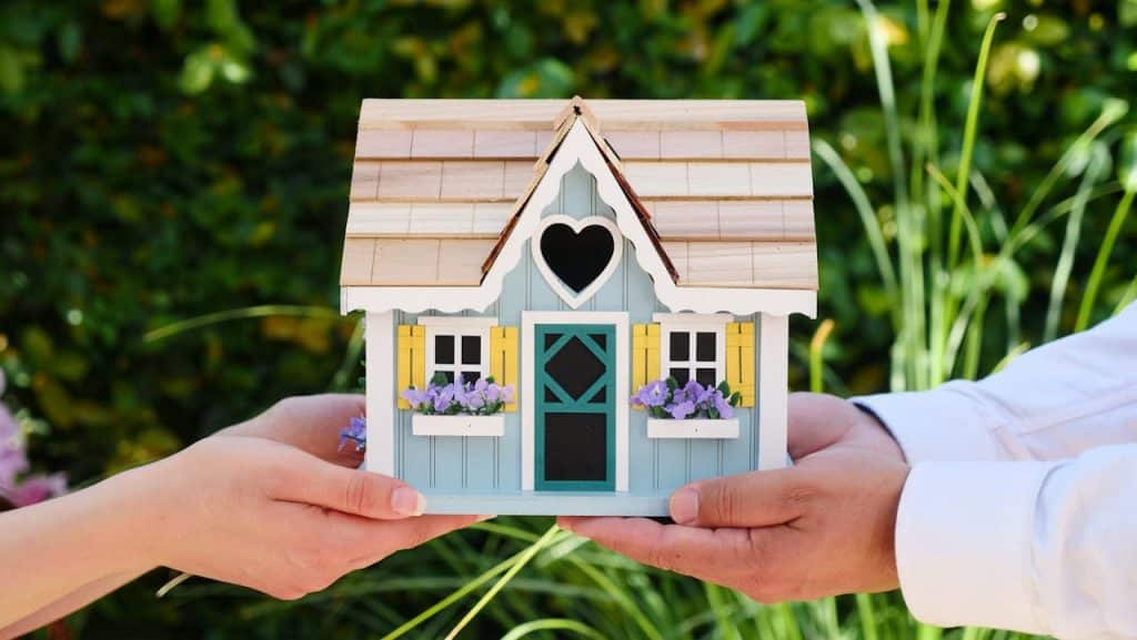 A pair of hands holding a charming miniature house with heart-shaped window and flower pots.