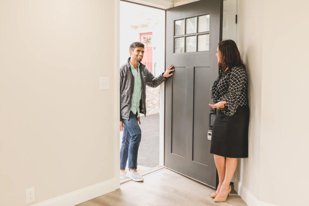 A man and woman stand together in front of a closed door, both smiling and dressed casually.