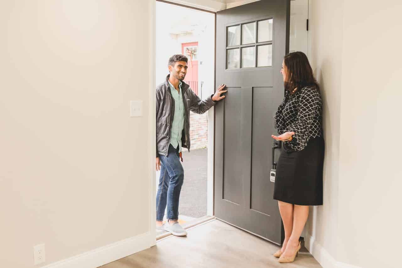 A man and woman stand together in front of a closed door, both smiling and dressed casually.