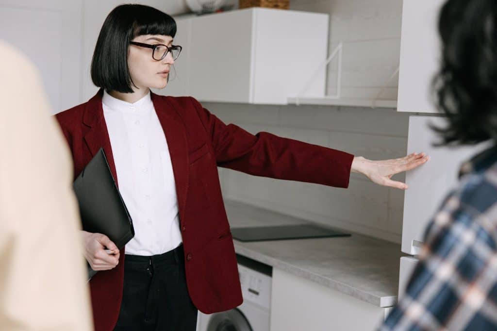 A woman wearing a red jacket and glasses stands in front of an open refrigerator, looking inside.