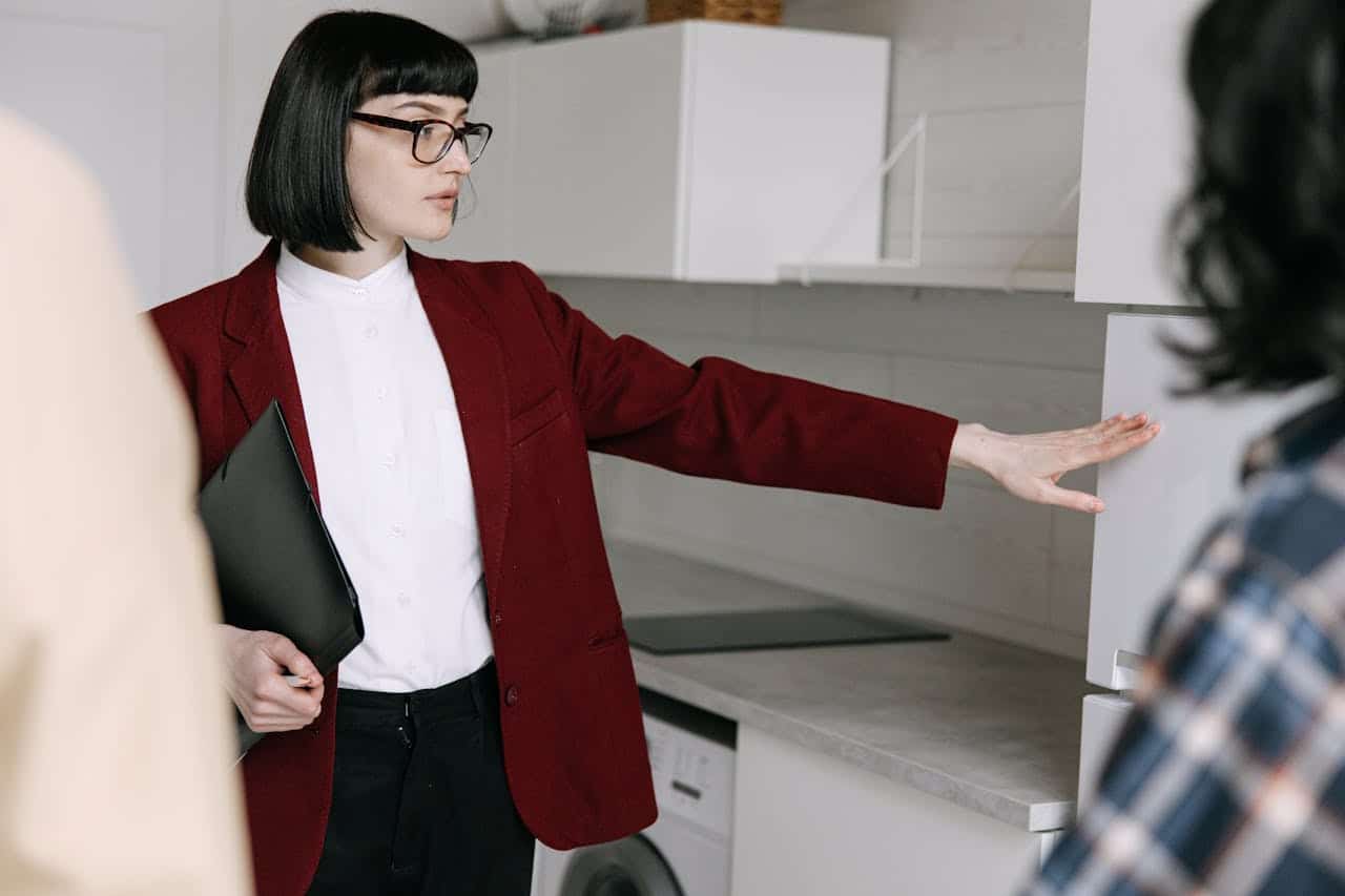 A woman wearing a red jacket and glasses stands in front of an open refrigerator, looking inside.