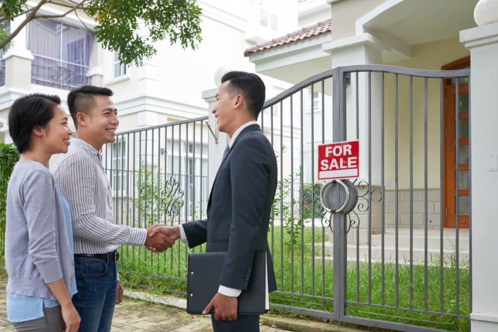 A couple shaking hands with a real estate agent in front of a house with a for sale sign.